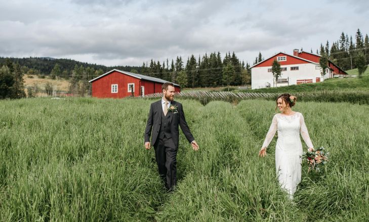 Newlyweds walking hand in hand through a lush green field near red barns on their wedding day.